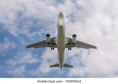 Commercial Passenger Plane Jet Aircraft From Below, Landing Gear Extracted And Landing Lights Switched On, Short Before Landing, Blue Sky And Clouds As Background