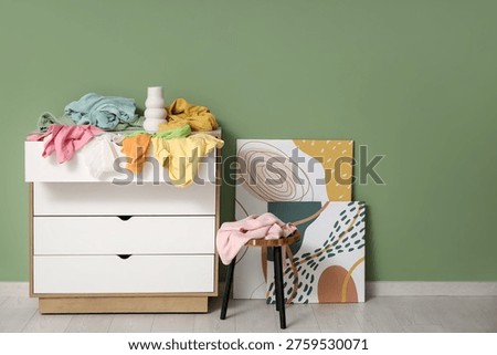 Commode with scattered clothes, table and paintings near green wall in room