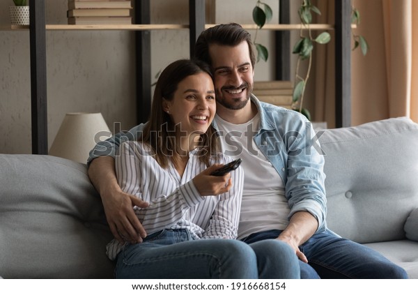 Common interests. Bonding millennial family\
couple hugging on sofa at living room having pleasure watching\
movie on tv. Friendly loving young spouses enjoy video film soccer\
match on weekend evening