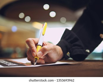 Common work practices theme hand holding a pen and writing signature for registration at seminar event, blurred background of hotel decorations in brown tones with bokeh defocused lights.