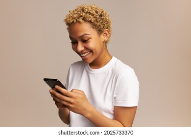 Communicating woman with Afro hairstyle using mobile phone, posing against wall with copy space for advertising content, having happy, cheerful look, reading funny message from her friend