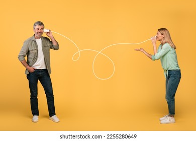 Communication and fun. Happy middle aged spouses talking through tin can telephone, standing over yellow studio background. Man and woman hearing and speaking through cups