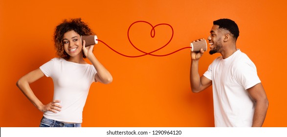 Communication And Fun. Young Black Couple Talking Through Tin Can Phone, Orange Background, Panorama