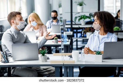 Communication and meeting in office after returning from covid-19 quarantine. Young guy and african american woman in protective mask talking through glass board at workplace with laptops in office - Powered by Shutterstock