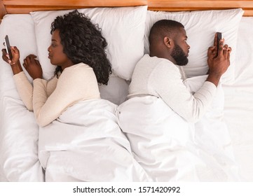 Communication and relationship problem. Black couple using smartphones in bed, lying back to back, ignoring each other after quarrel