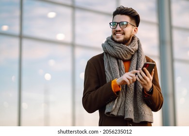 Оnline communication. Young man wearing  coat with a smartphone in his hand, walking in the street. Young bearded guy with modern hairstyle in urban background  typing on cell phone.  - Powered by Shutterstock