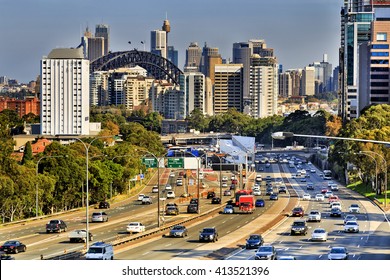 Communing Car Traffic During Rush Hour In Busy City Centre Of Sydney. Multi Lane Motorway Between Towers Of Modern Architecture Skyscrapers With Sydney Harbour Bridge In The Background.