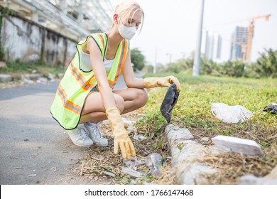 Community Service - Young Female Volunteer Person Cleaning The Littered Streets In A Urban Environment By Picking Up Trash Laying On The Ground While Wearing A Face Mask And A Reflective Vest.