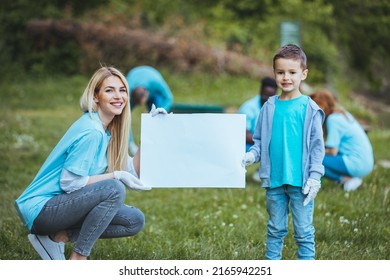 Community Volunteers Are Cleaning Their Local Park. The Family Recycles Garbage. A Mother Stands In The Foreground With Her Son And Holds A Sign While Other Family Members Collect Garbage In The Back