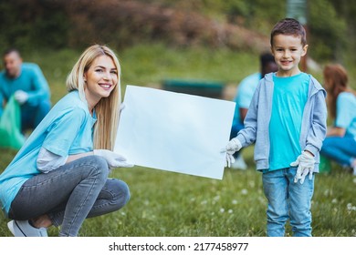 Community Volunteers Are Cleaning Their Local Park. The Family Recycles Garbage. A Mother Stands In The Foreground With Her Son And Holds A Sign While Other Family Members Collect Garbage