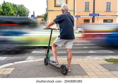 Commuter Hipster Man Using Electric Scooter Is Waiting To Cross A Road With Heavy Traffic. Eco Transport Concept 