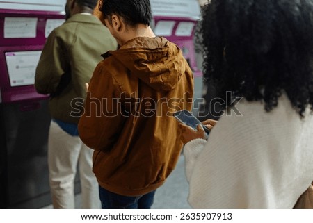 Commuters using smart phones while waiting to buy tickets at a modern ticket vending machine in a subway station