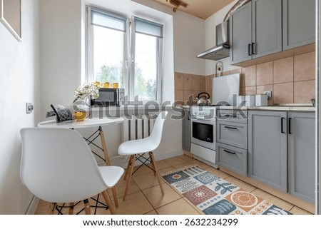 A compact bright kitchen corner features a round white table with chairs, a floral vase and microwave by a window above gray cabinets and stove.
