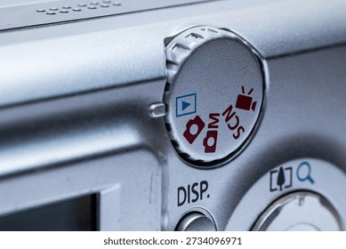 Compact digital camera mode selection dial showing photo, video, and playback icons.