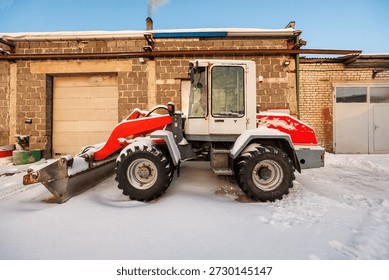 Compact front end loader with red arm and white cab sits by brick and cinder block walls, tires and bucket edged with fresh snow, warm low light, chimney exhaust visible.