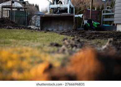 A compact front loader digs a narrow trench in a suburban backyard at dusk, bucket mid scoop, tire tracks and soil clods visible near fences, sheds, and a ride on toy.