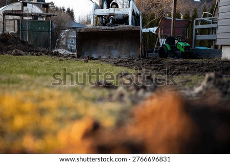 A compact front loader digs a narrow trench in a suburban backyard at dusk, bucket mid scoop, tire tracks and soil clods visible near fences, sheds, and a ride on toy.