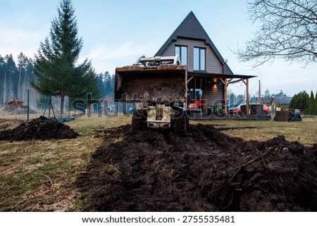 A compact front loader sits in a muddy trench facing a dark wood A frame cabin with porch, yard tools, and a small green tractor, conifers, wire fence, and outbuildings.