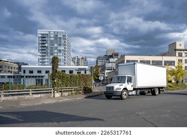 Compact Industrial spacious white middle class relocation rig day cab semi truck tractor with long box trailer running for delivery on the urban city road with apartment and office buildings