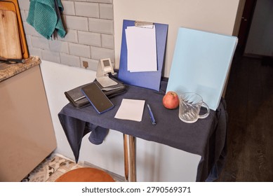 Compact kitchen table setup displaying smartphone, clipboard with blank paper, blue folder, notepad, pen, red apple, and empty glass cup against white tiled wall and wooden floor background