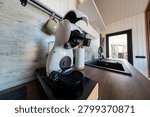 A compact kitchenette centers on a capsule espresso machine with head raised on a dark wooden counter beside a double sink and gooseneck faucet, early day natural light.
