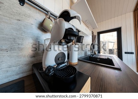 A compact kitchenette centers on a capsule espresso machine with head raised on a dark wooden counter beside a double sink and gooseneck faucet, early day natural light.