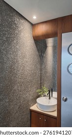 A compact powder room is elevated through a strong focus on texture, contrast, and restraint. The dominant element is the rough, tactile wall finish—its irregular surface catching light in subtle gradients, creating depth within a small footprint.

The clean geometry of the white vessel sink provides a deliberate contrast, acting as a visual pause against the textured backdrop. Paired with a matte black faucet, the composition gains a modern, refined edge without feeling sterile.

Warm wood cabinetry and framing soften the space, introducing balance and continuity with the rest of the interior language. The curved transition at the upper cabinet detail is particularly notable—it breaks rigidity and adds a crafted, almost bespoke quality.

A mirrored surface extends the spatial perception, making the room feel larger while doubling the textural richness of the wall. The soft overhead lighting enhances this effect, emphasizing material rather than form.

The frosted glass door with circular accents introduces a subtle retro-industrial touch, tying back to the broader design narrative seen in previous spaces.

Despite its limited size, the space feels intentional and complete—each material plays a role, and nothing feels excessive.
