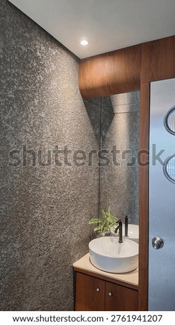 A compact powder room is elevated through a strong focus on texture, contrast, and restraint. The dominant element is the rough, tactile wall finish—its irregular surface catching light in subtle gradients, creating depth within a small footprint.

The clean geometry of the white vessel sink provides a deliberate contrast, acting as a visual pause against the textured backdrop. Paired with a matte black faucet, the composition gains a modern, refined edge without feeling sterile.

Warm wood cabinetry and framing soften the space, introducing balance and continuity with the rest of the interior language. The curved transition at the upper cabinet detail is particularly notable—it breaks rigidity and adds a crafted, almost bespoke quality.

A mirrored surface extends the spatial perception, making the room feel larger while doubling the textural richness of the wall. The soft overhead lighting enhances this effect, emphasizing material rather than form.

The frosted glass door with circular accents introduces a subtle retro-industrial touch, tying back to the broader design narrative seen in previous spaces.

Despite its limited size, the space feels intentional and complete—each material plays a role, and nothing feels excessive.
