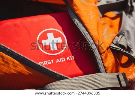 Compact Red First Aid Kit Visible, Closeup Of Zippered Pouch Inside Orange Rucksack, White Cross Emblem Clear, Textured Fabric With Water Beads, Trailready Medical Supply For Hiker Emergency Care