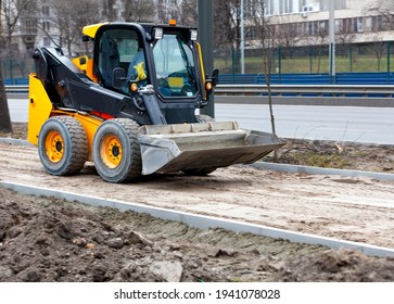 Compact road grader on a construction site is leveling a sandy base on a sidewalk path under construction. Light blur in motion, city road background.