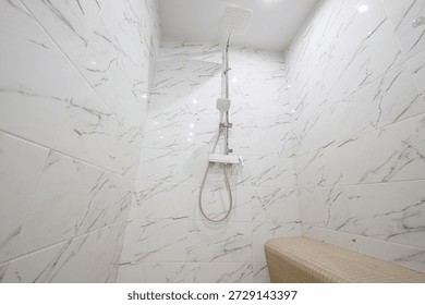  A compact shower cabin with white marble-patterned tiles, a fixed showerhead, and a built-in wooden bench. Clean, minimalist design with recessed ceiling lighting.
