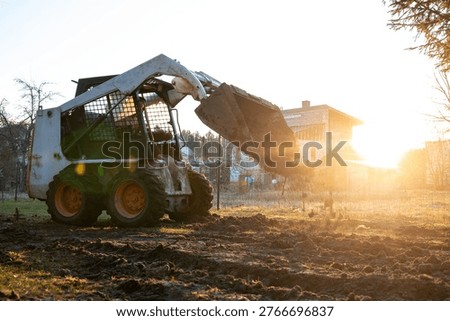 A compact skid steer with caged cab dumps soil on a muddy plot at sunrise near a partially built brick structure with open window bays, shot low with lens flare.