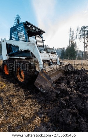 A compact skid steer loader digs wet soil, orange wheels caked in mud, boxy cab with metal grates, leafless trees and open field, low angle, early day light.