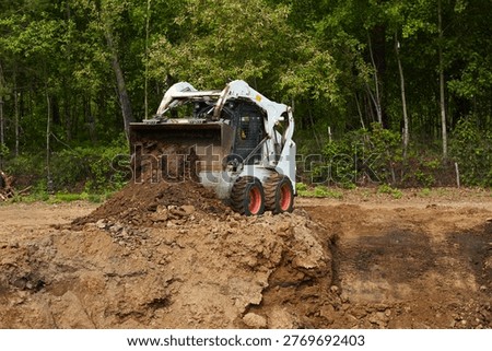 Compact skid steer loader pushing and dumping soil on a construction site near a forest, shaping terrain for future building work.