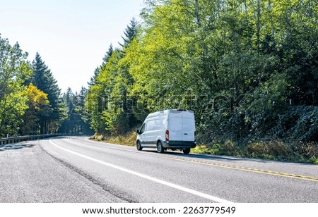 Compact small size white mini van with cargo compartment for local deliveries and small business needs for customers commercial cargo delivery driving on the turning road with forest on the sides