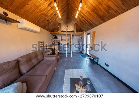 Compact tiny house interior with loft, ladder, and warm pendant lights. Vaulted plank ceiling, kitchenette, brown sectional, wall AC, and coffee table suggest a retreat.