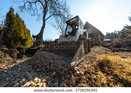 A compact track loader dumps crushed stone and levels a muddy driveway beside a wooden house, with a leafless tree and tall evergreens under bright mid day light.