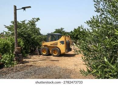 A compact yellow skid steer loader parked on a dirt ground surrounded by green trees and agricultural fields under a clear blue sky at a local rural farm attraction during a sunny day in Israel