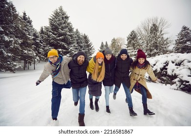 Company of cheerful young women and men have fun together on vacation in winter forest. Smiling friends in winter casual clothes embracing running on snow looking at camera. Friends and winter concept