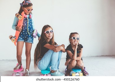 Company of children of different ages wearing cool fashion clothing posing with colorful skateboards against white wall, urban style