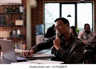 Company employee with physical disability talking on landline phone call, using office telephone at desk. Wheelchair user having remote conversation on line, dealing with impairment. - Powered by Shutterstock