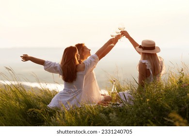 The company of female friends enjoys a summer picnic and raise glasses with wine. Happy women in the park in evening sunlight. The lights of a sun.  - Powered by Shutterstock