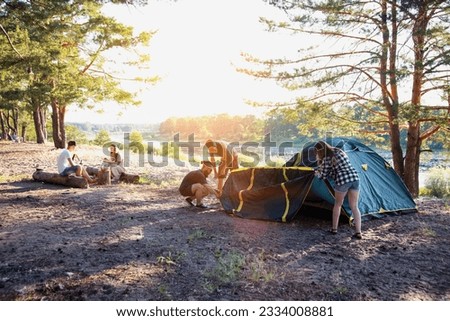 A company of friends guys and girls set up a tent on the sandy bank of the river in summer. Hiking, outdoor recreation in the forest. Sunny day, countryside