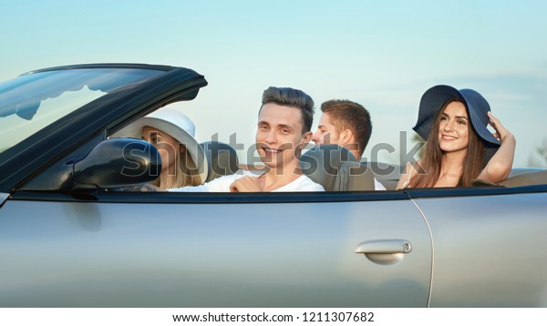 Company going for drive in silver cabriolet,\
couples sitting in automobile. Young gorgeous girls wearing in brim\
broad hats and two handsome men traveling by car. Concept of\
summertime, adventure.