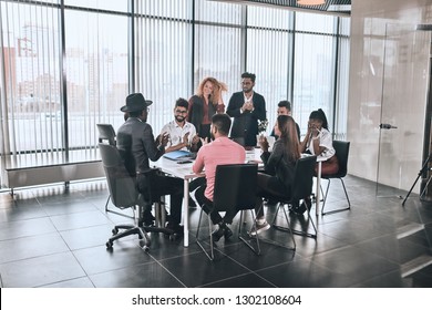 Company Is Greeting Foreign Partner. Happy Diverse People Are Sitting At The Table And Clapping Hands In The Room With Panorama Window