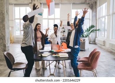 Company of happy multiracial coworkers wearing casual outfits standing around table with documents and raised hands while looking at each other and celebrating achievement - Shutterstock ID 2201646229