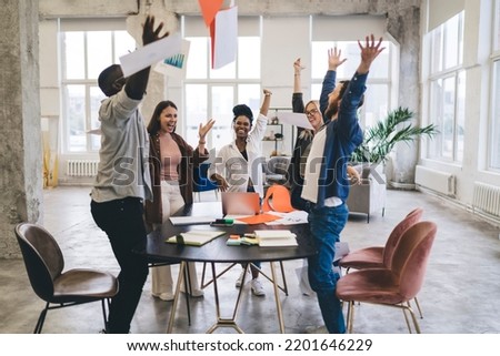 Company of happy multiracial coworkers wearing casual outfits standing around table with documents and raised hands while looking at each other and celebrating achievement