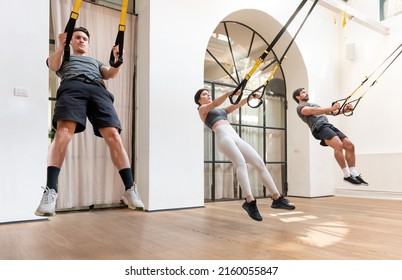 Company Of Male And Female Athletes Jumping And Doing Squat Exercise With TRX Ropes During Active Workout In Gym