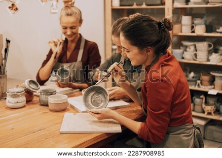 A company of three cheerful young women friends are painting ceramics in a pottery workshop. Have fun doing art.