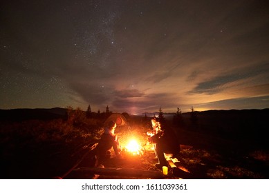 Company Of Three Tourists Travelers Men And Woman Sitting By Burning Campfire On Grassy Mountain Valley With Small Spruce Trees, Enjoying Beautiful Camping Night Under Dark Starry Cloudy Sky.